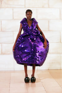 Model in a voluminous purple sequin gown posing against a pale stone wall.