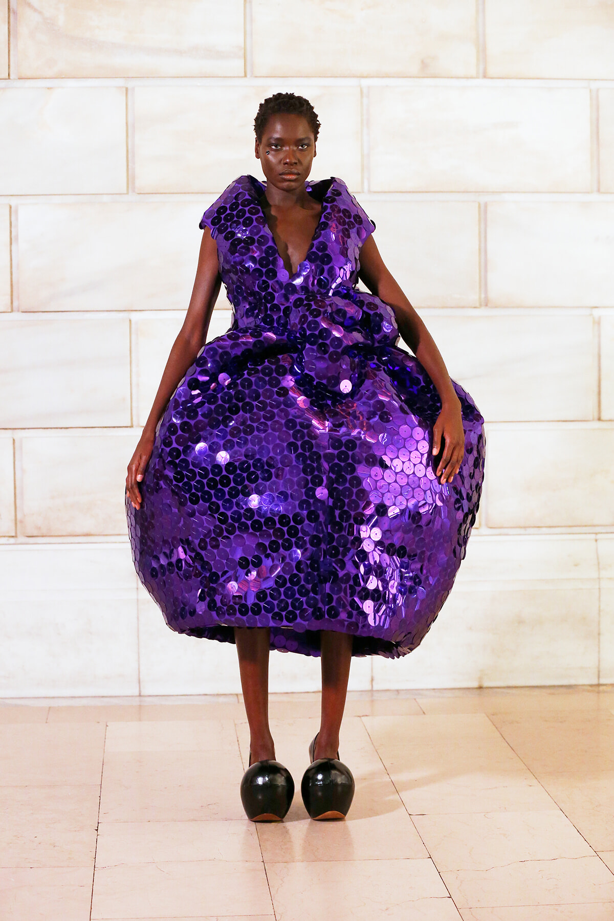 Model in a voluminous purple sequin gown posing against a pale stone wall.