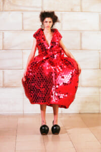 Model posing in a voluminous red sequined dress with circular disc embellishments against a light stone wall.