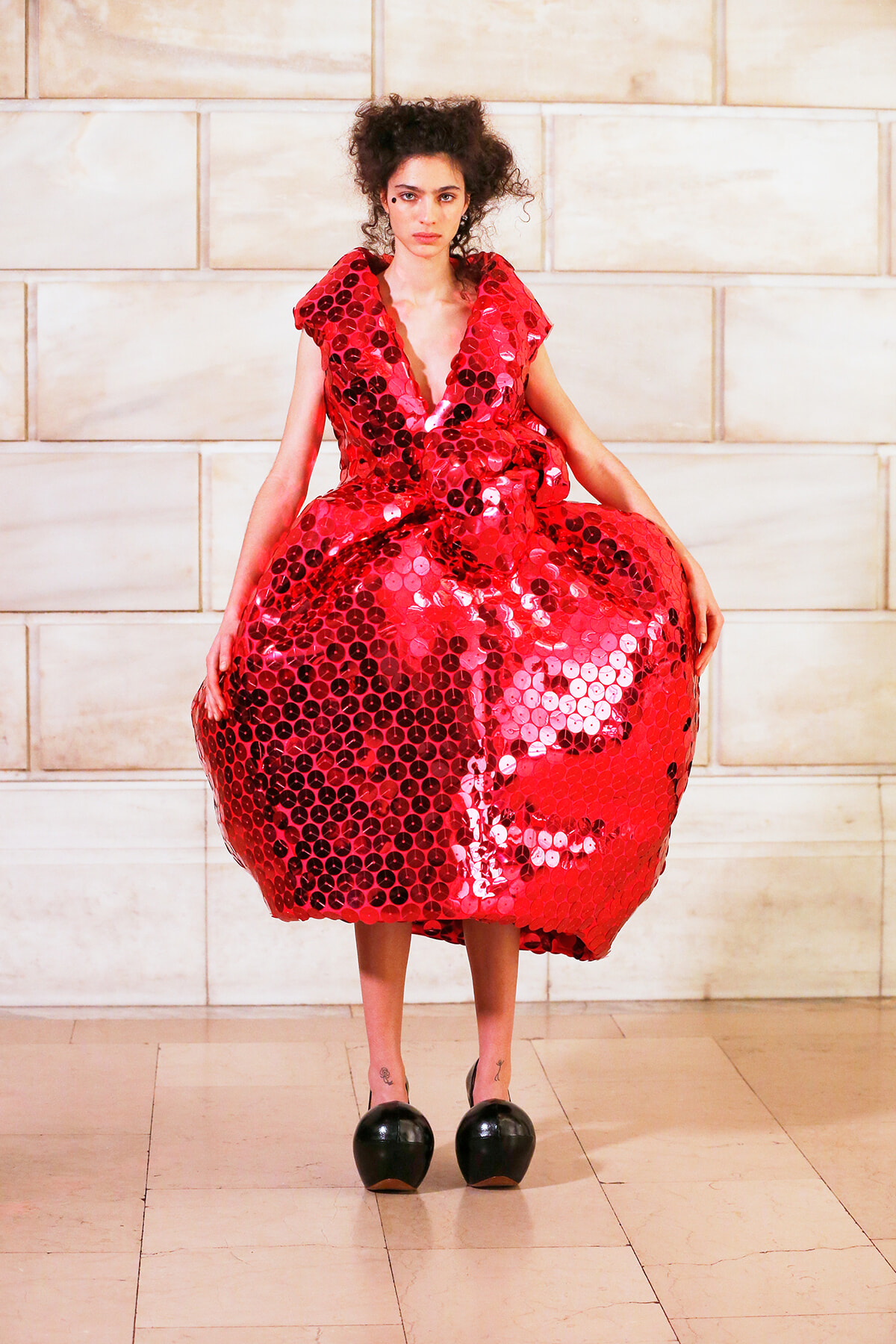 Model posing in a voluminous red sequined dress with circular disc embellishments against a light stone wall.