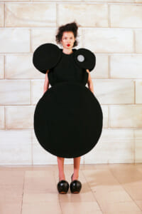 Model wearing an avant-garde black dress with oversized circular shoulder and hip shapes, standing against a light stone wall.