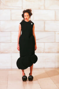 Model in a black sleeveless sheath dress with large circular fabric circles at the hem, standing indoors against a light stone wall.