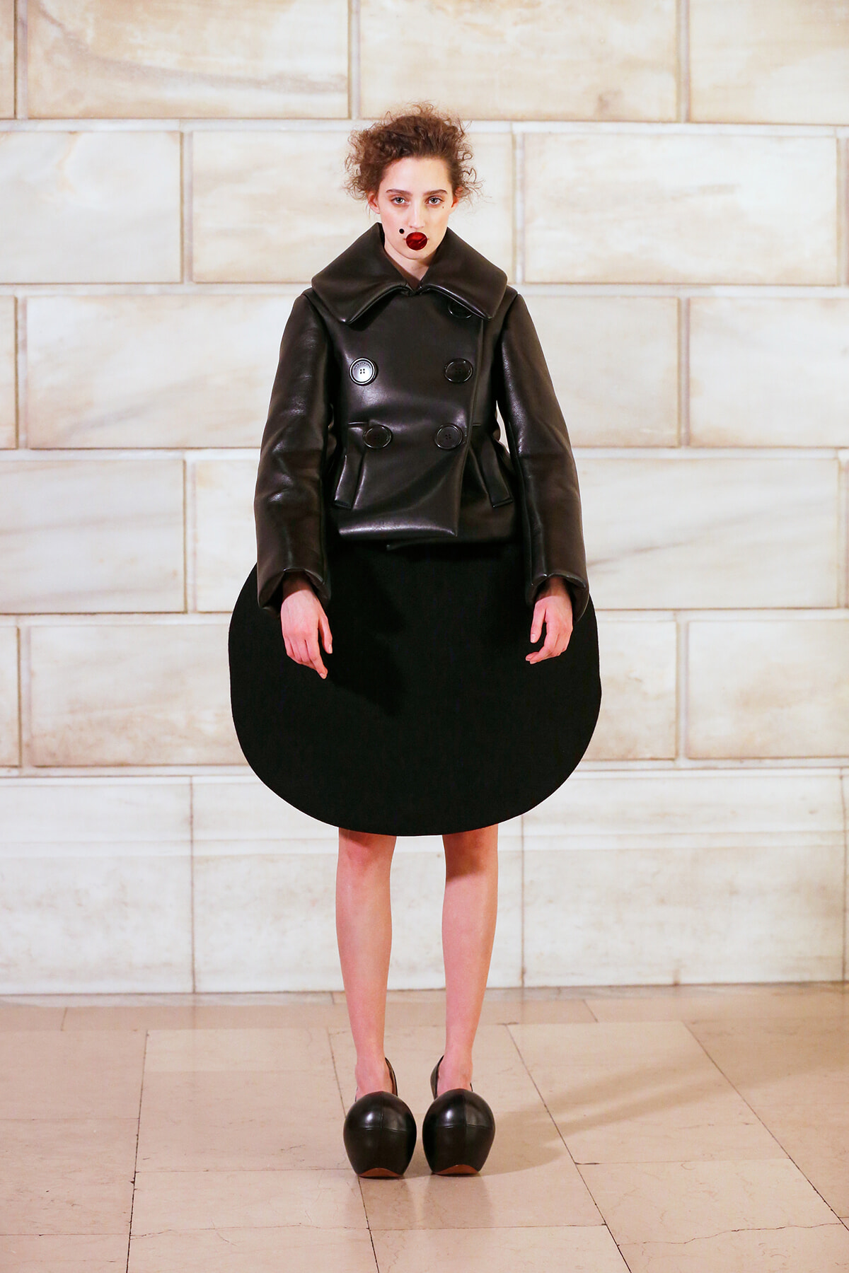 Model wearing a black leather double-breasted coat with a large rounded black skirt, standing against a pale stone wall.