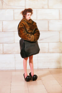 Young woman in a chunky brown fur sweater, oversized black bag, and black platform heels stands against a stone wall.