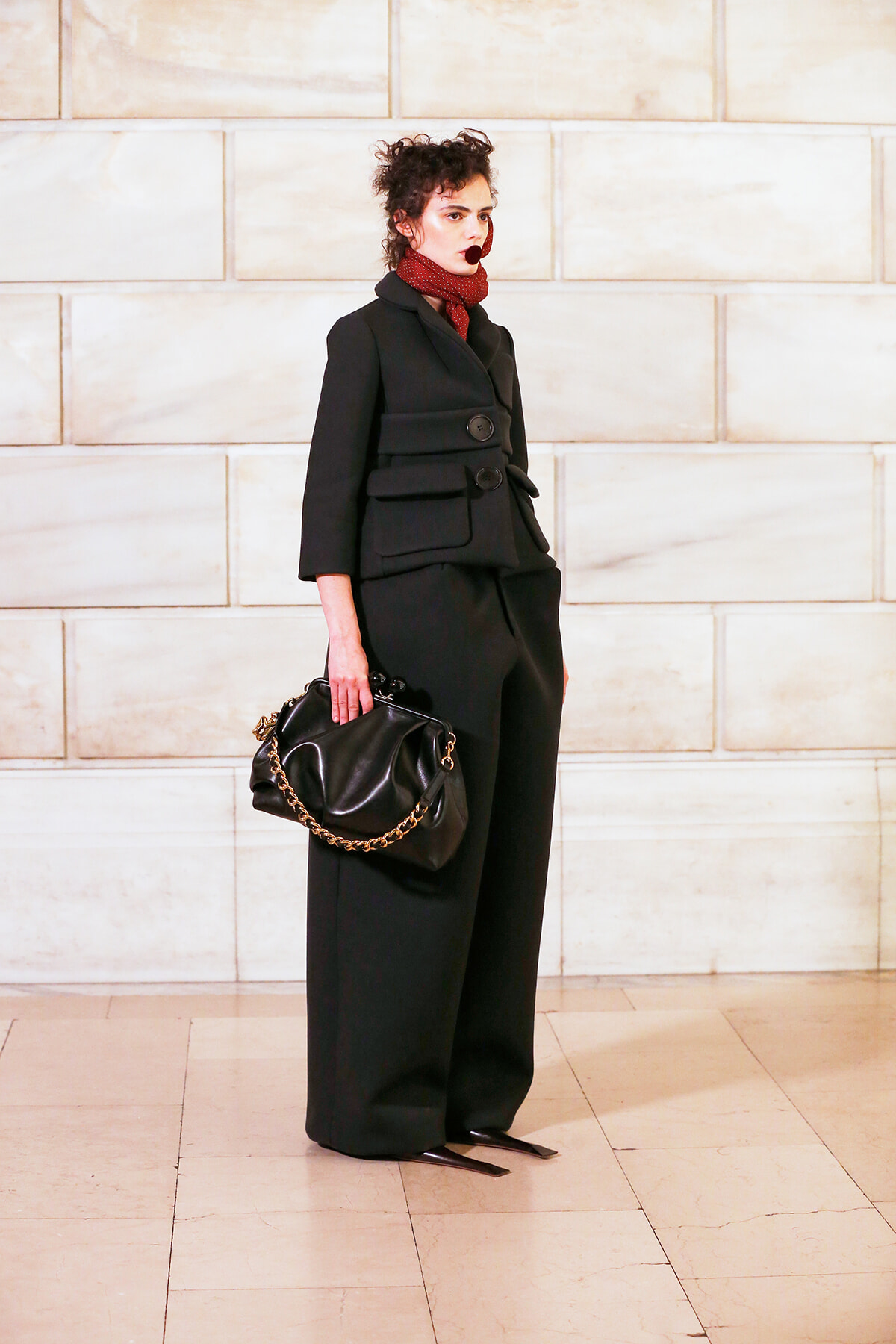 Individual wearing a black tailored double-breasted suit with a red scarf, holding a black handbag with a gold chain against a pale stone wall.