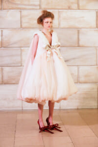 Model in a pale pink, voluminous dress with a large satin bow at the waist, standing against a stone wall.
