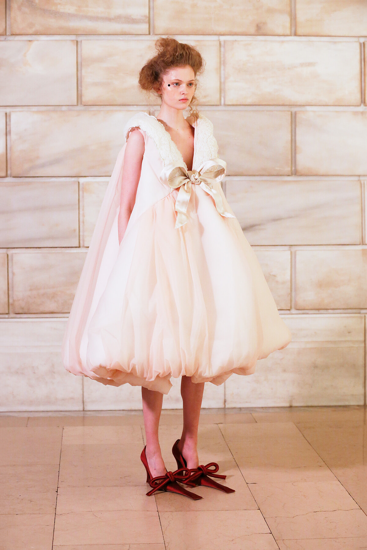 Model in a pale pink, voluminous dress with a large satin bow at the waist, standing against a stone wall.