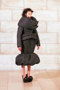 Model wearing an avant-garde, oversized dark gray coat with a large wraparound collar and a rounded, bubble-shaped skirt, standing against a pale stone wall.