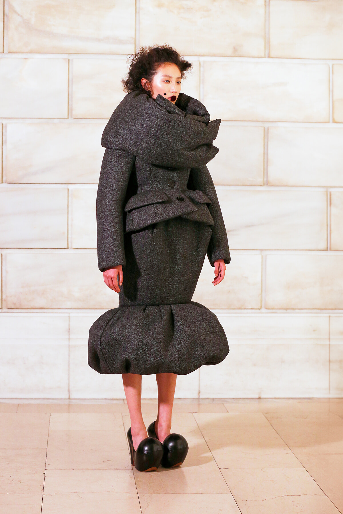 Model wearing an avant-garde, oversized dark gray coat with a large wraparound collar and a rounded, bubble-shaped skirt, standing against a pale stone wall.
