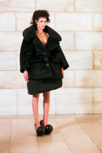 Model wearing an oversized black coat with a large collar and buttons, standing against a beige stone wall.