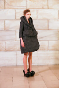 Model in a dark checkered oversized coat with large padded pockets, standing in a light marble setting; wearing extreme black platform shoes with rounded balls at the toes.