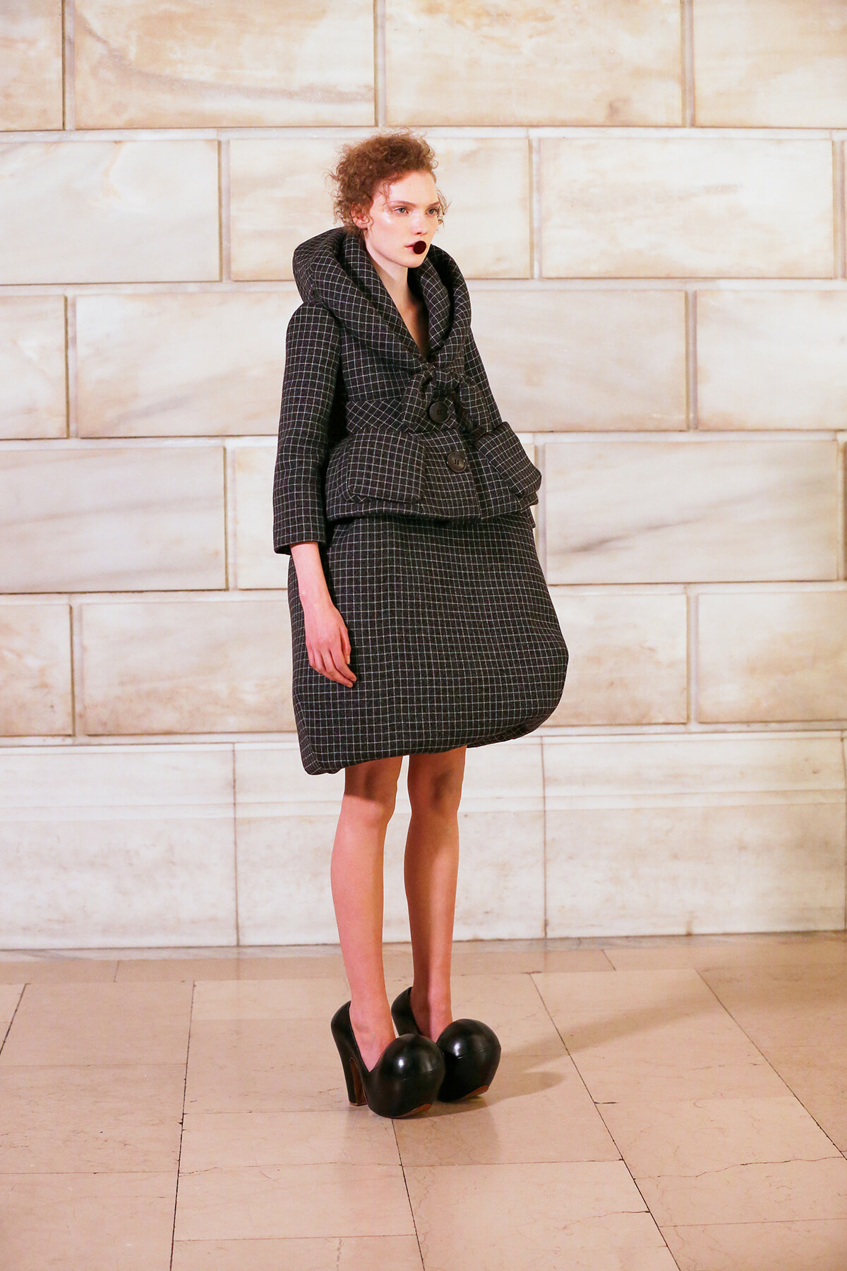 Model in a dark checkered oversized coat with large padded pockets, standing in a light marble setting; wearing extreme black platform shoes with rounded balls at the toes.