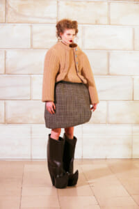 A model in a beige chunky cardigan over a matching sweater and plaid knee-length skirt, wearing tall black heeled boots against a light stone wall backdrop.