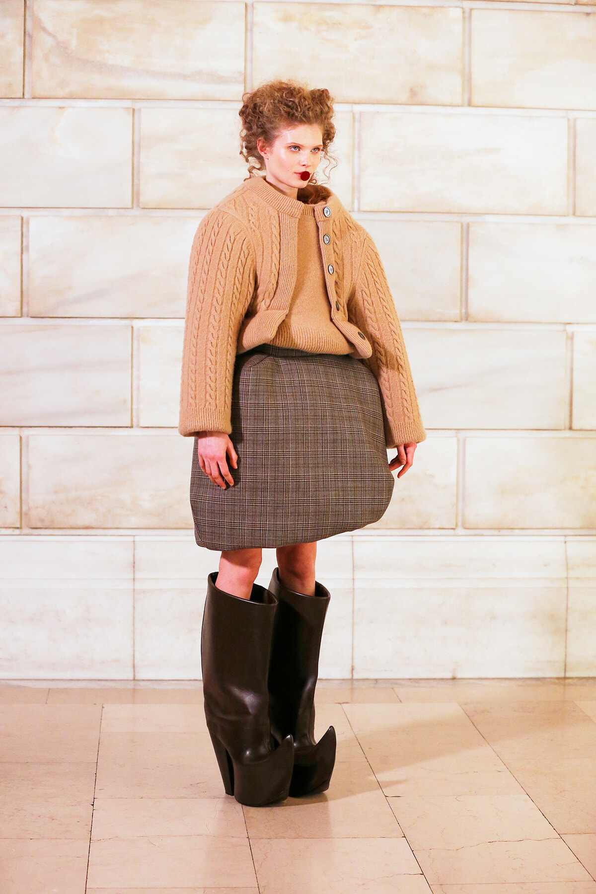 A model in a beige chunky cardigan over a matching sweater and plaid knee-length skirt, wearing tall black heeled boots against a light stone wall backdrop.