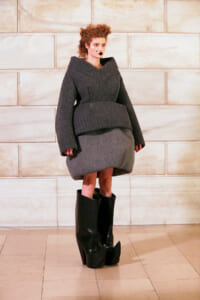 Model in a chunky gray knit sweater with an oversized peplum, gray pencil skirt, and tall black platform boots on a light tiled floor.