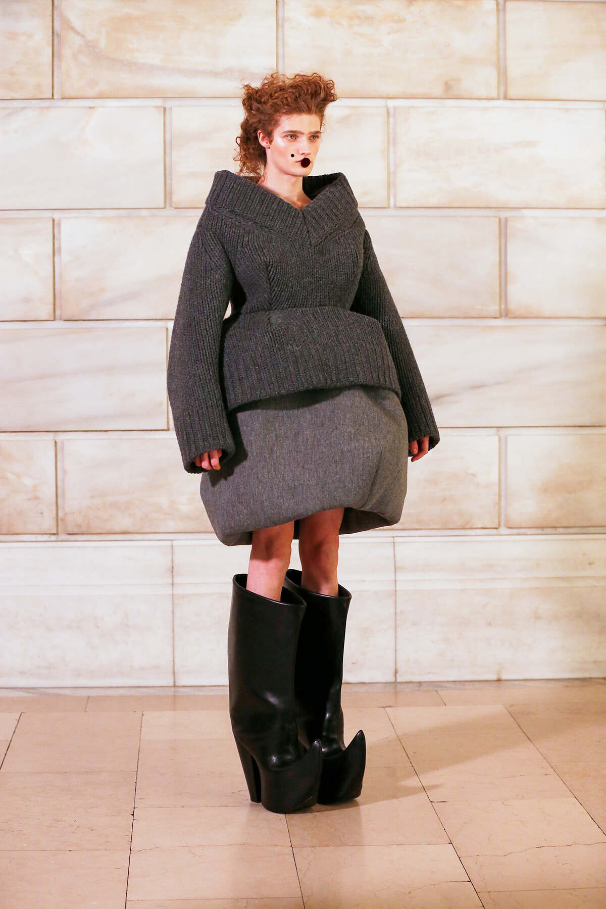 Model in a chunky gray knit sweater with an oversized peplum, gray pencil skirt, and tall black platform boots on a light tiled floor.