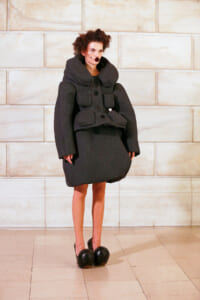 Model wearing an oversized dark gray coat with a large collar and front pockets, standing against a light stone wall.