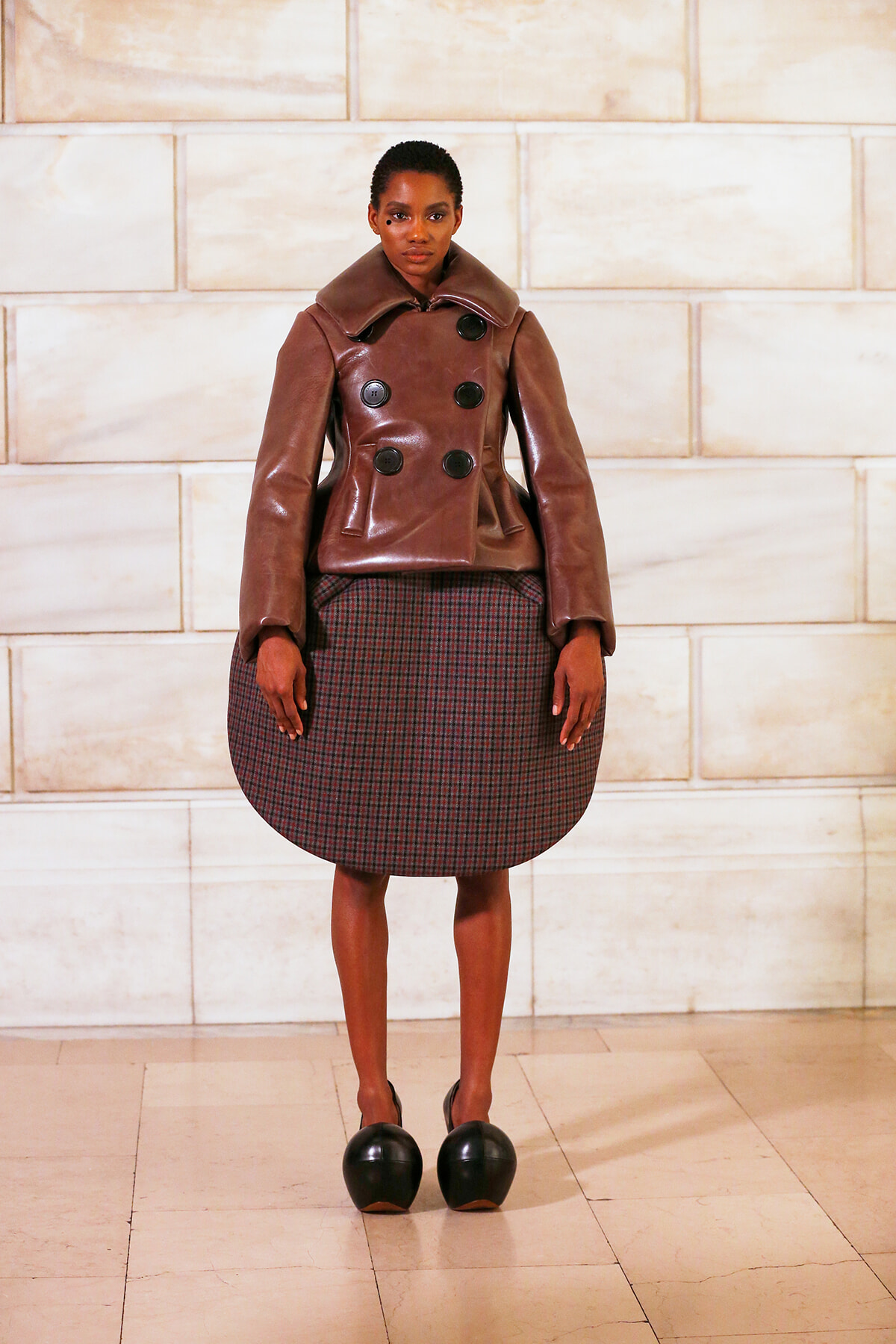 Female model on a fashion runway wearing a brown leather double-breasted coat with large black buttons, paired with a rounded plaid skirt and black clogs against a light stone wall backdrop.