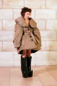Model wearing an oversized beige puffer coat with a huge collar, belt, and knee-high black platform boots against a light stone wall.