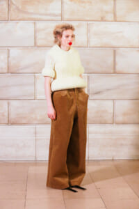 Woman in an oversized cream knit sweater and wide-leg brown corduroy pants stands against a beige stone wall.