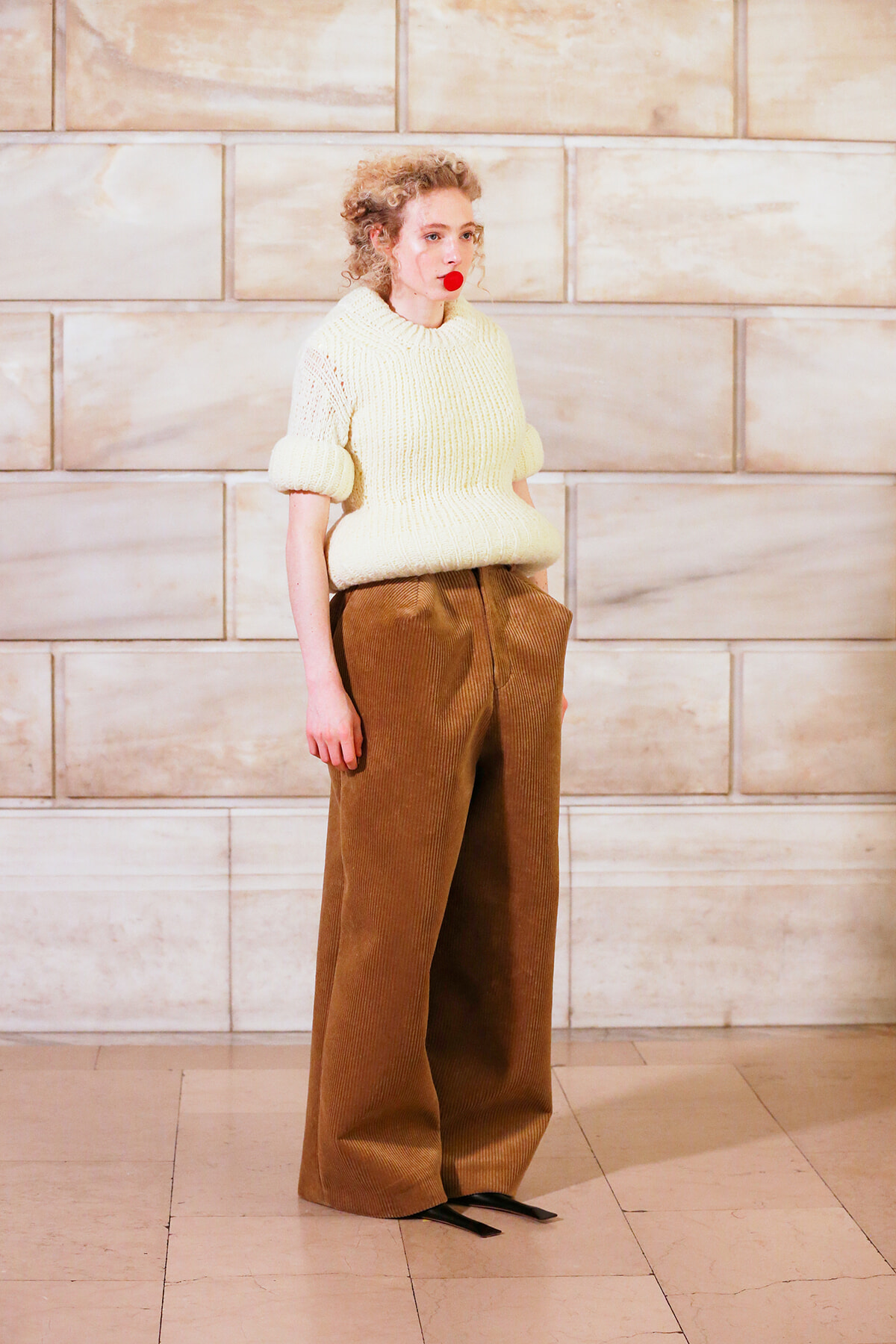 Woman in an oversized cream knit sweater and wide-leg brown corduroy pants stands against a beige stone wall.