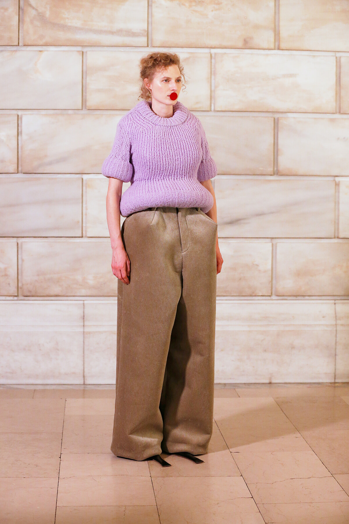 Person wearing a chunky lavender knit sweater and wide-leg beige corduroy pants stands against a light stone wall.