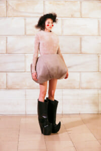 Model wearing a pale pink velvet dress with an oversized bubble skirt and knee-high black boots against a light stone wall background
