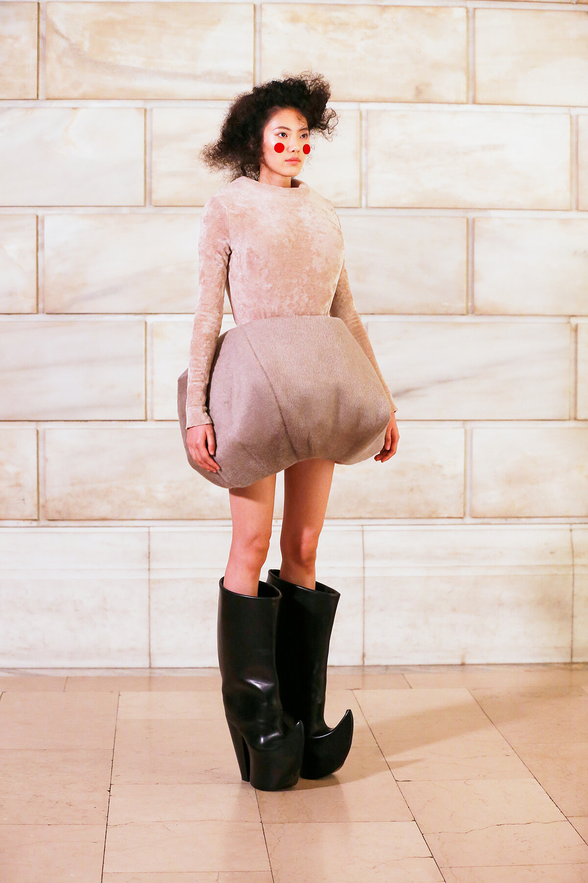 Model wearing a pale pink velvet dress with an oversized bubble skirt and knee-high black boots against a light stone wall background
