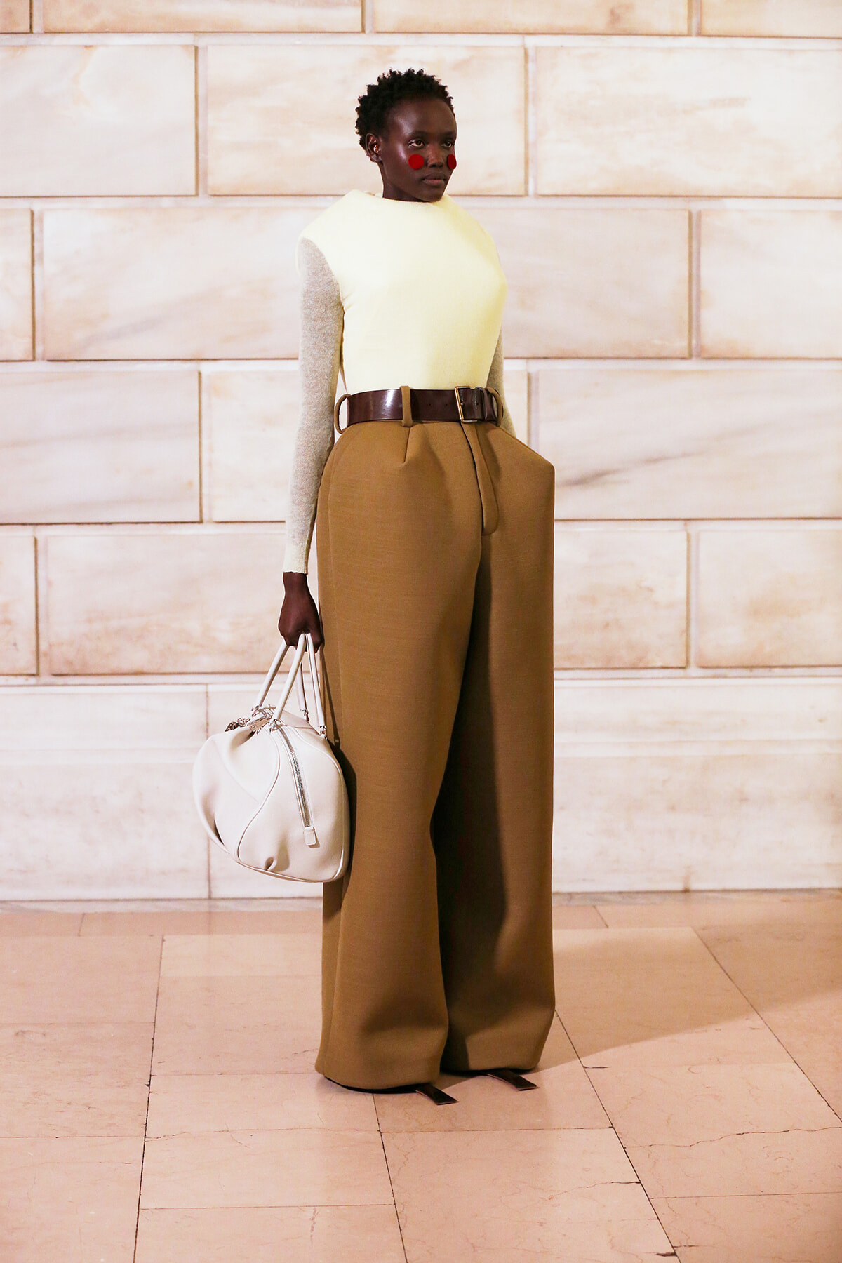 Model wearing a pale yellow top with beige sleeves tucked into high-waisted camel wide-leg trousers, accessorized with a wide brown belt and a white handbag, standing against a light stone wall.