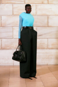 Model wearing a turquoise sweater with black wide-leg pants and a thick belt, holding a black handbag, posed against a light stone wall.