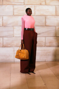 Model stands against a stone wall in a pink knit top, burgundy wide-leg pants with a black belt, and a tan handbag in hand.