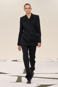 Model walking a fashion runway in a black tailored pantsuit with a wrap blazer and matching trousers.