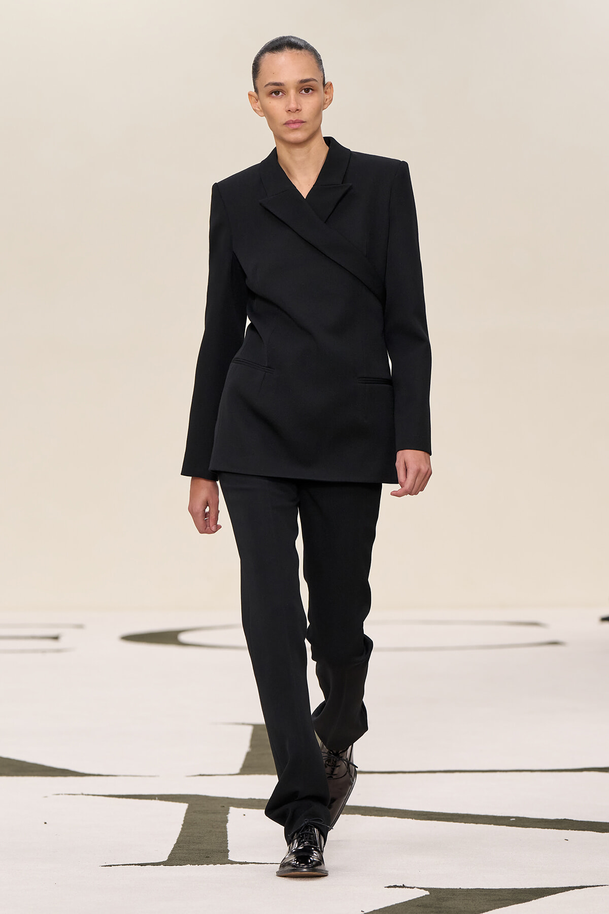 Model walking a fashion runway in a black tailored pantsuit with a wrap blazer and matching trousers.