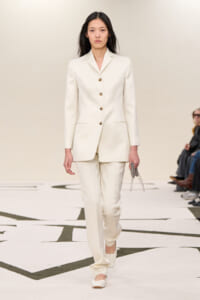 Model wearing a cream tailored blazer and matching trousers walking a runway, holding a small chain handbag.