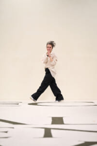 Person in beige shirt and black wide-leg pants walks while hands are pressed together in a prayer-like pose against a pale backdrop.