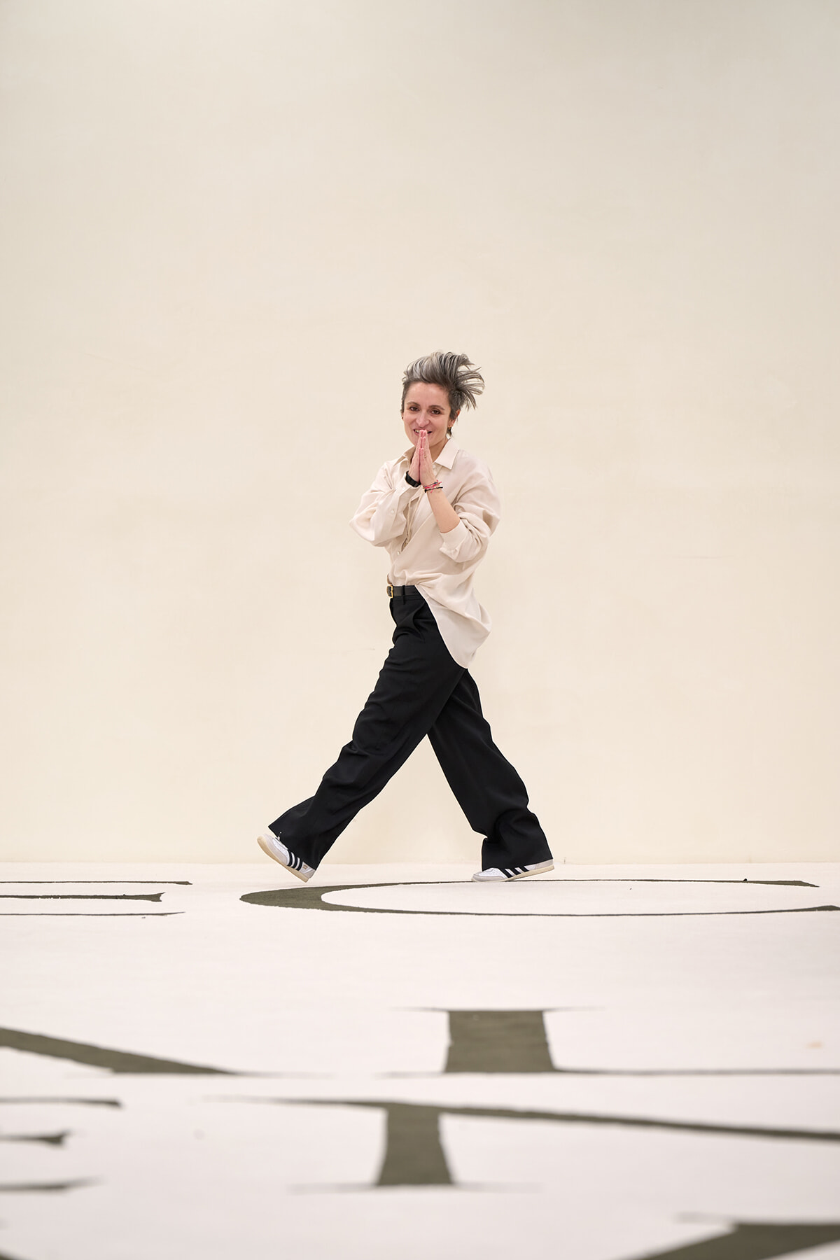 Person in beige shirt and black wide-leg pants walks while hands are pressed together in a prayer-like pose against a pale backdrop.