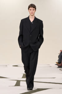 Male model in a tailored black suit walking the runway with hands in pockets.