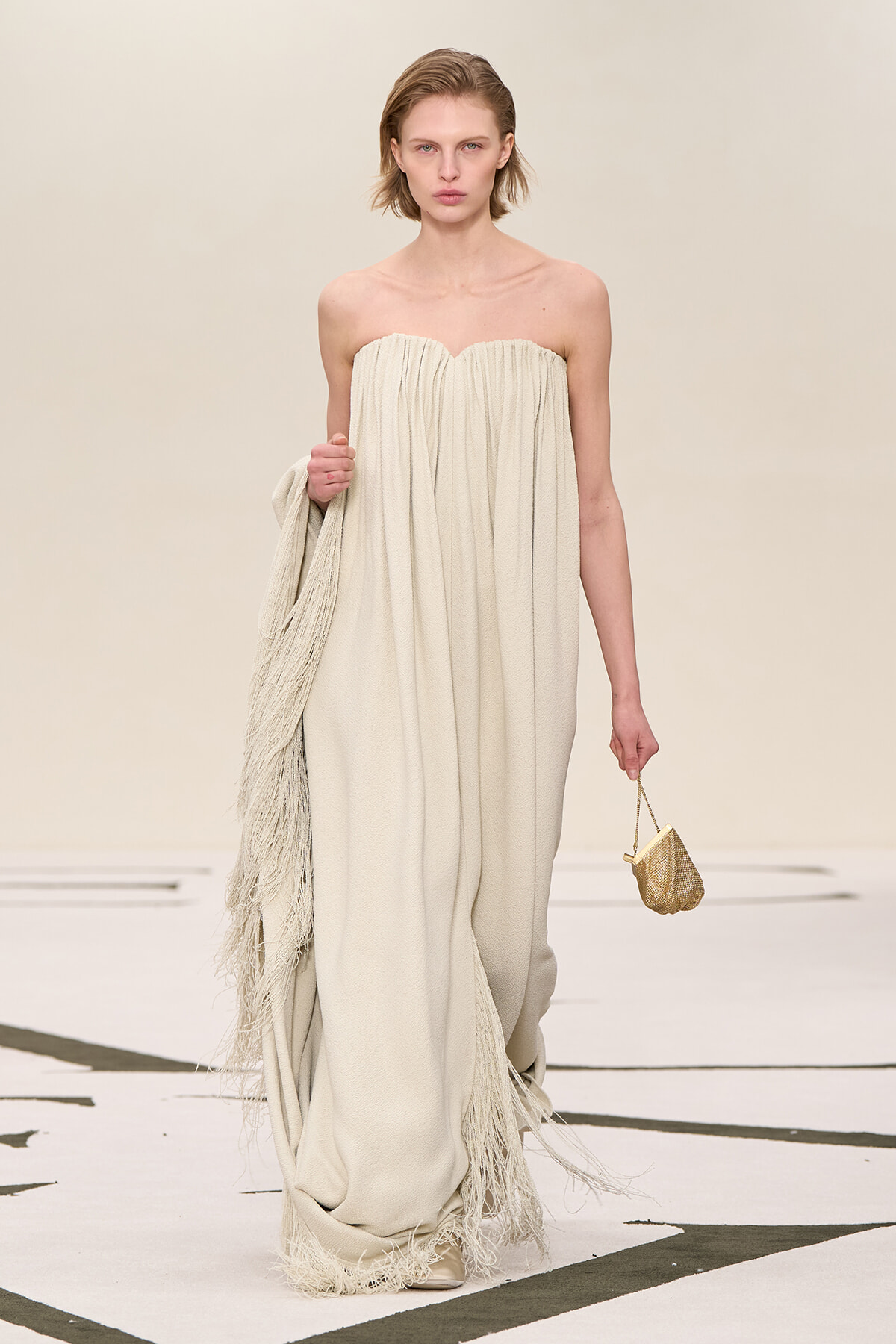 Model on a runway wearing a strapless, floor-length cream gown with fringe trim, holding a matching fringed wrap and a small gold handbag.