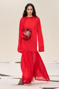 Model walks a runway wearing a bright red long-sleeve gown with a draped sheer hem, holding a red clutch.