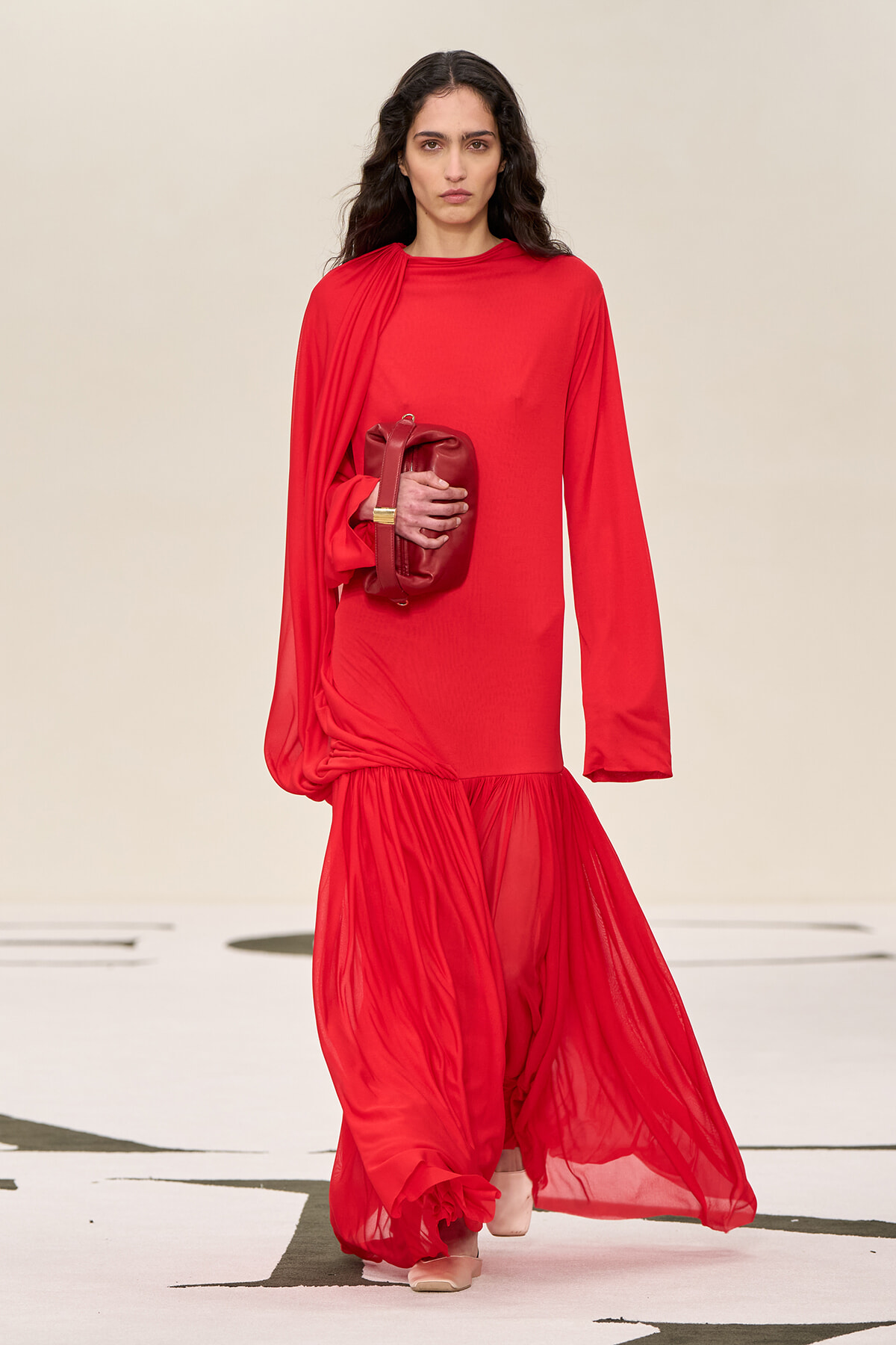 Model walks a runway wearing a bright red long-sleeve gown with a draped sheer hem, holding a red clutch.