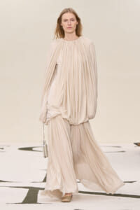 Runway model wearing a soft beige, draped, long-sleeve gown with a gathered neckline and a small beige handbag walking on a white catwalk.