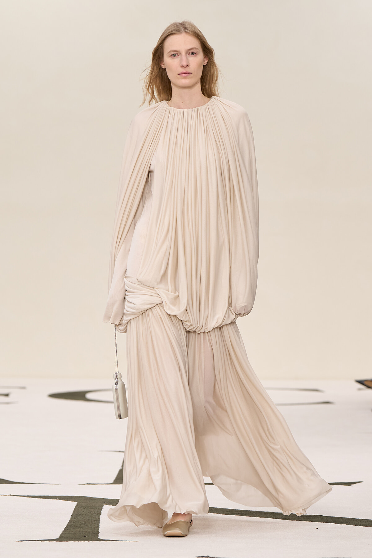 Runway model wearing a soft beige, draped, long-sleeve gown with a gathered neckline and a small beige handbag walking on a white catwalk.