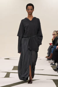 Model walks a fashion runway wearing an oversized black hoodie top with a gathered draped skirt and dark flats, accessorized with glasses