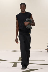 Male model walks the runway in a sleeveless black top and loose black pants, carrying a gray leather bag tucked under his arm.