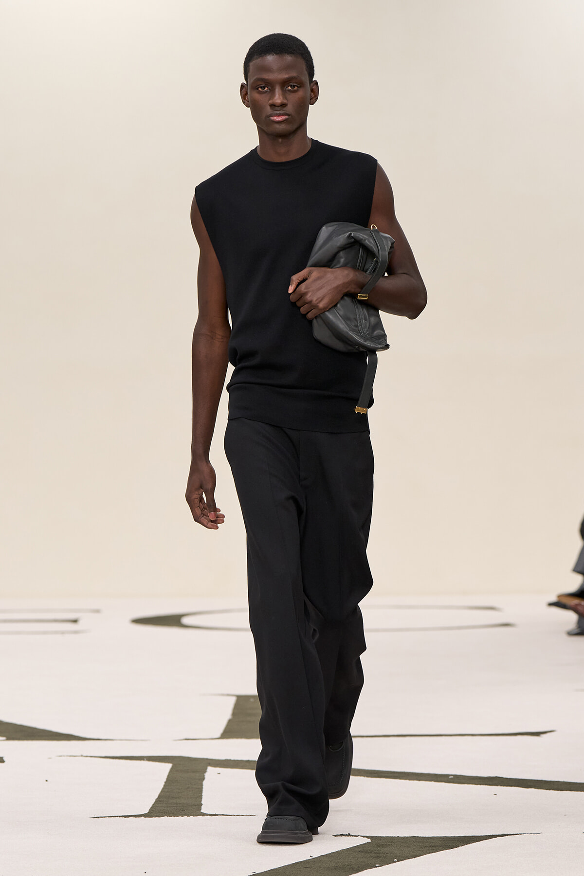 Male model walks the runway in a sleeveless black top and loose black pants, carrying a gray leather bag tucked under his arm.