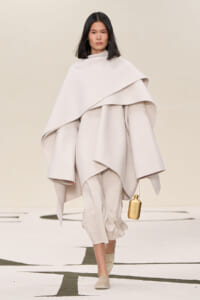 Model walking in a pale cream oversized cape and layered outfit, carrying a small gold handbag.
