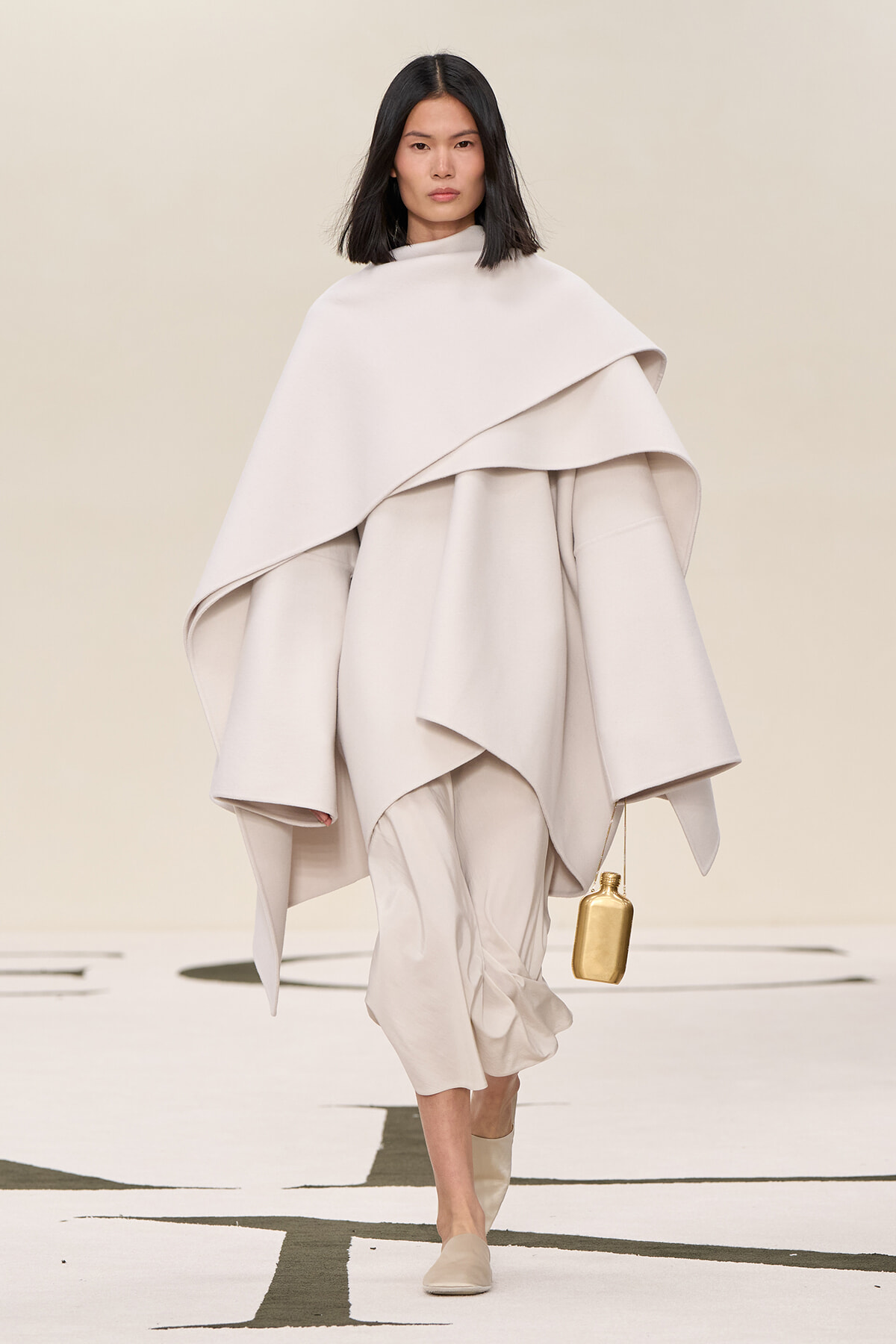 Model walking in a pale cream oversized cape and layered outfit, carrying a small gold handbag.
