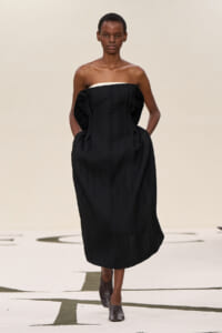 Model walking a runway in a strapless black pleated midi dress with white neckline edge and pockets, paired with brown ankle boots.
