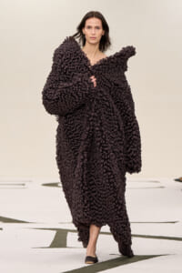 Model walks the runway wearing an oversized dark brown loop-knit cape with voluminous sleeves.