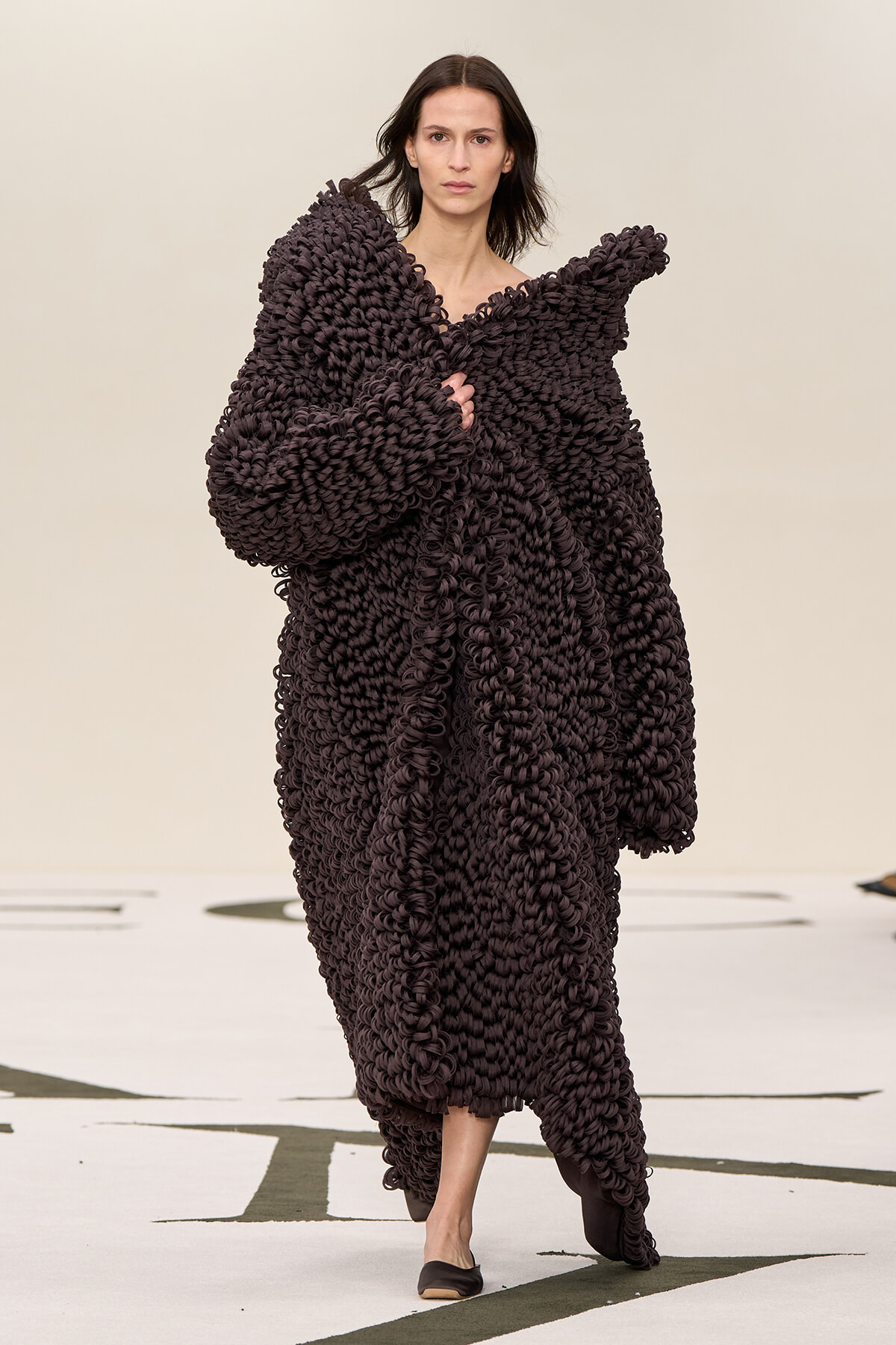 Model walks the runway wearing an oversized dark brown loop-knit cape with voluminous sleeves.